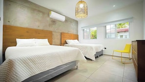 Une chambre d’hôtel avec deux lits, une tête de lit en bois, un appareil de climatisation, une chaise jaune et une fenêtre donnant sur un paysage verdoyant.