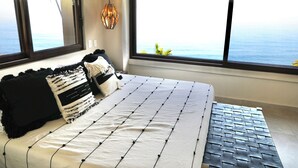 Ein Schlafzimmer mit einem Bett, Kissen, einer Hängeleuchte und großen Fenstern mit Meerblick.