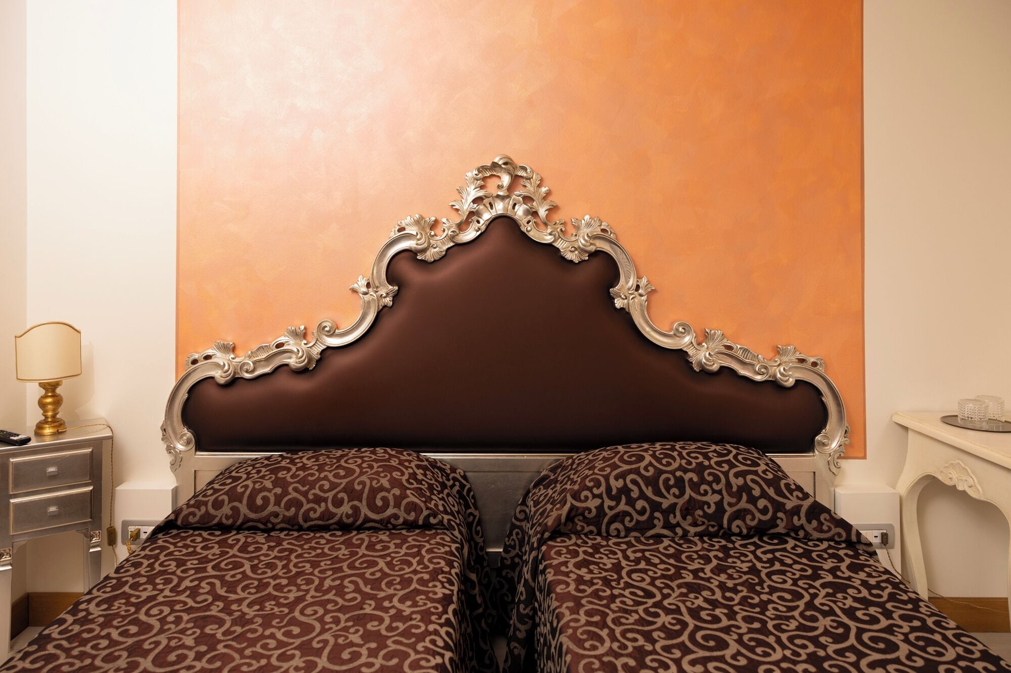 A bedroom with a large ornate headboard, two patterned beds, a nightstand with a lamp, and a small table with decorative items.