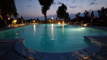 A swimming pool at dusk with surrounding lounge chairs and umbrellas, surrounded by trees and a clear sky.