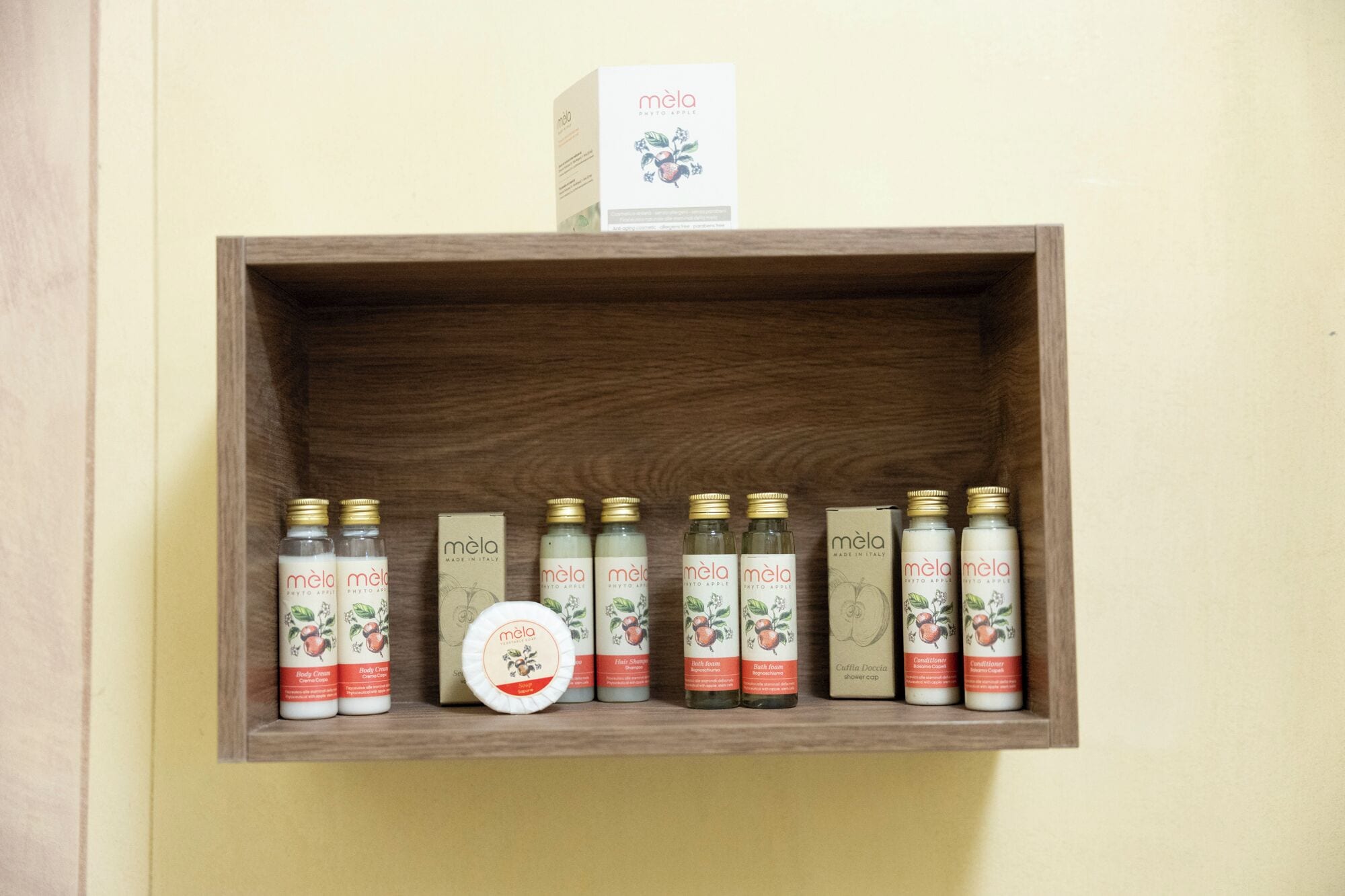 Un estante de madera con varios productos Mela, incluyendo crema corporal, champú para cabello, espuma de baño, gorro de ducha y acondicionador.