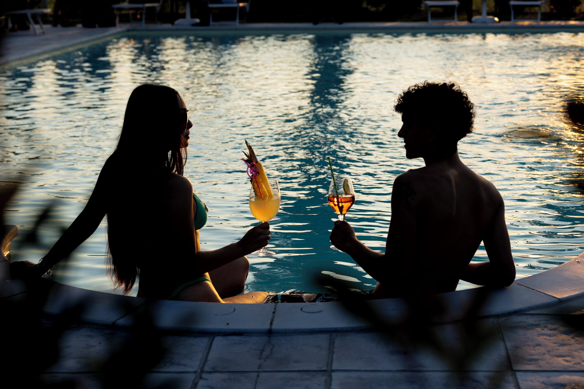 Dos personas sentadas junto a una piscina con bebidas, rodeadas de sillones de descanso y un suelo de baldosas.