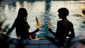 Dos personas sentadas junto a una piscina con bebidas, rodeadas de sillones de descanso y un suelo de baldosas.
