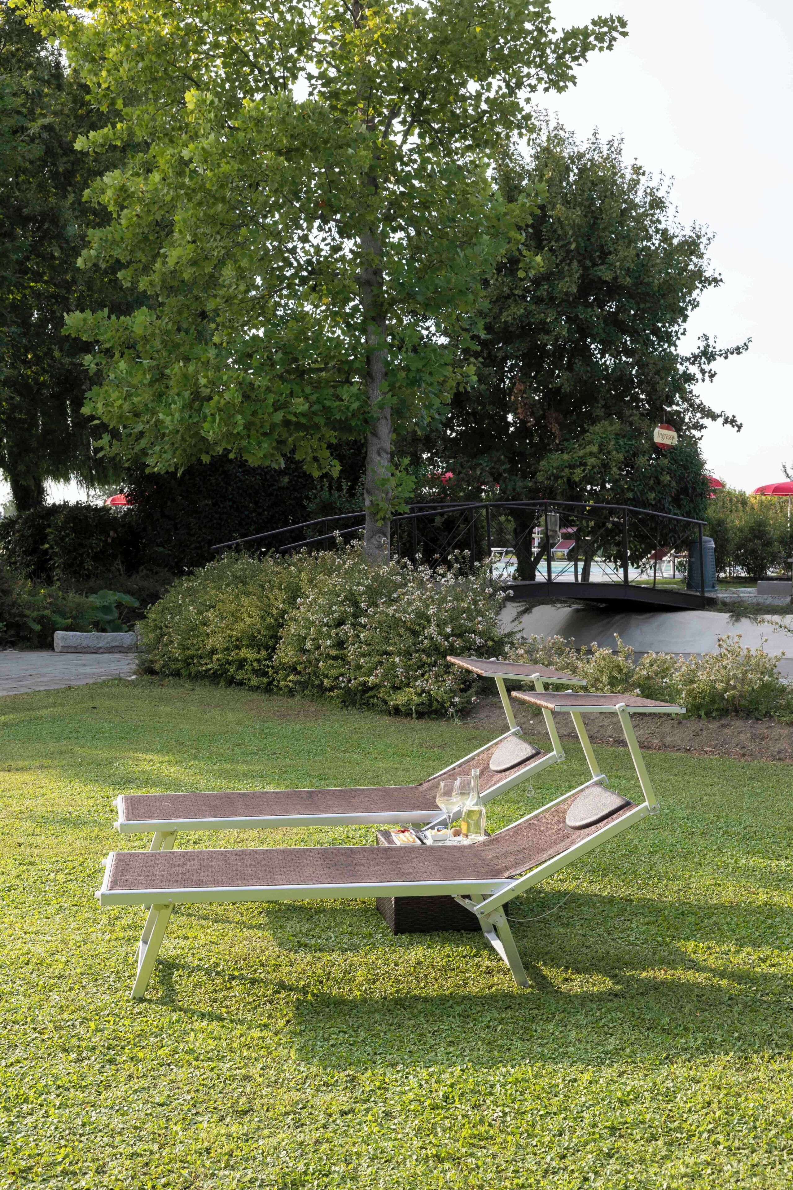 Two sun loungers with cushions and a glass of wine on a grassy area with a bridge and trees in the background.
