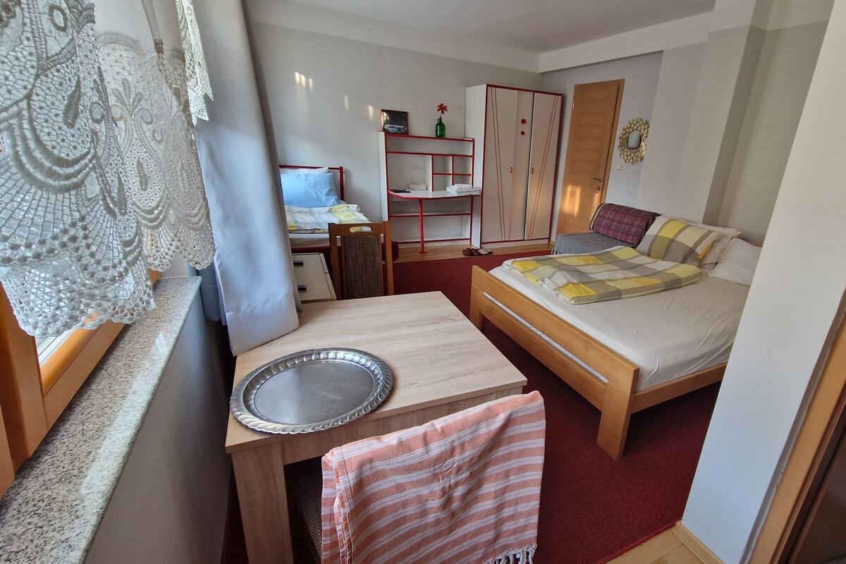 A compact bedroom with a single bed, a small desk, a wardrobe, and a window with lace curtains.