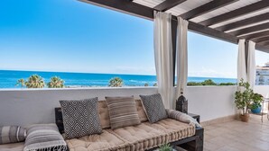 A balcony with a cushioned seating area, a small table with decorative pots, and a view of the ocean. - Apartment in Playas del Duque for 5 people | Casa Cadiz 412A (Puerto Banús - Marbella)