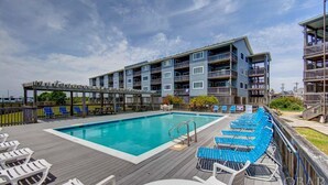 A swimming pool with lounge chairs, a wooden deck, and a multi-story building with balconies. - New Listing-Open weeks in prime summer time! Cozy condo, ocean views & pool.   (Rodanthe)