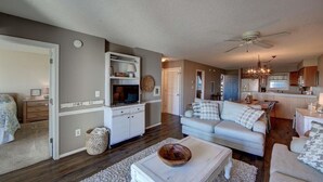 A spacious living room with a sofa, coffee table, and a television. There is a kitchen area with cabinets and a dining table. - New Listing-Open weeks in prime summer time! Cozy condo, ocean views & pool.   (Rodanthe)