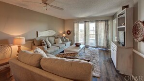 A living room with a sofa, coffee table, and a ceiling fan. There is a window with curtains and a view outside. - New Listing-Open weeks in prime summer time! Cozy condo, ocean views & pool.   (Rodanthe)