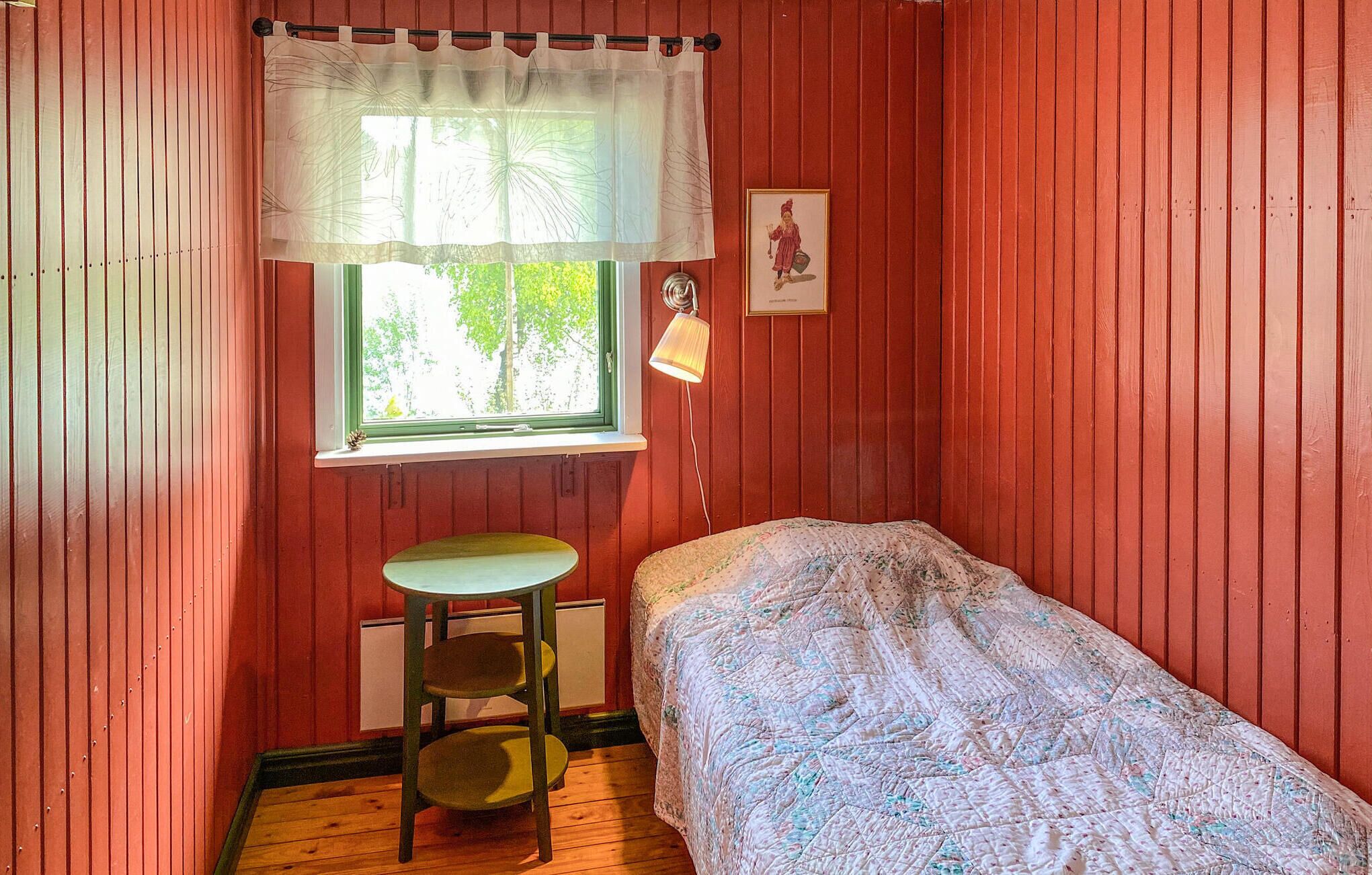 A small room with a single bed, a window with a white curtain, a small table with a stool, and a wall-mounted lamp.