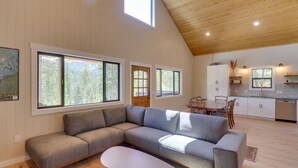 Uma sala de estar moderna com sofá seccional, mesa de jantar e área de cozinha com armários brancos e teto de madeira.