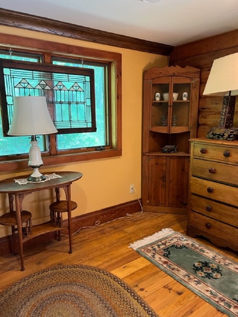 Ein Zimmer mit Holzboden, einem Tisch mit Lampe, einem Schrank mit Glastüren und einem gemusterten Teppich.