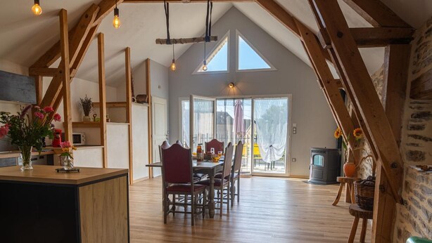 A spacious loft with high ceilings, exposed wooden beams, a dining area with a table and chairs, and a large sliding glass door leading to an outdoor patio. - Gîte La Bergerie 5/6 Pers (LACHAMP-RIBENNES)