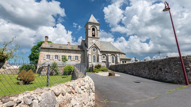 A historic stone church with a bell tower, a stone wall, and a paved road leading to it.