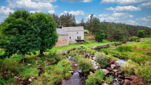 A stone house with a balcony, surrounded by lush greenery and a small stream. - Le Moulin de Jeannou (PEYRE EN AUBRAC)