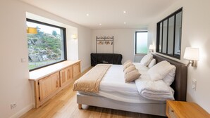 A modern bedroom with a large bed, a wooden bench at the window, and a view of greenery outside. - Le Moulin de Jeannou (PEYRE EN AUBRAC)