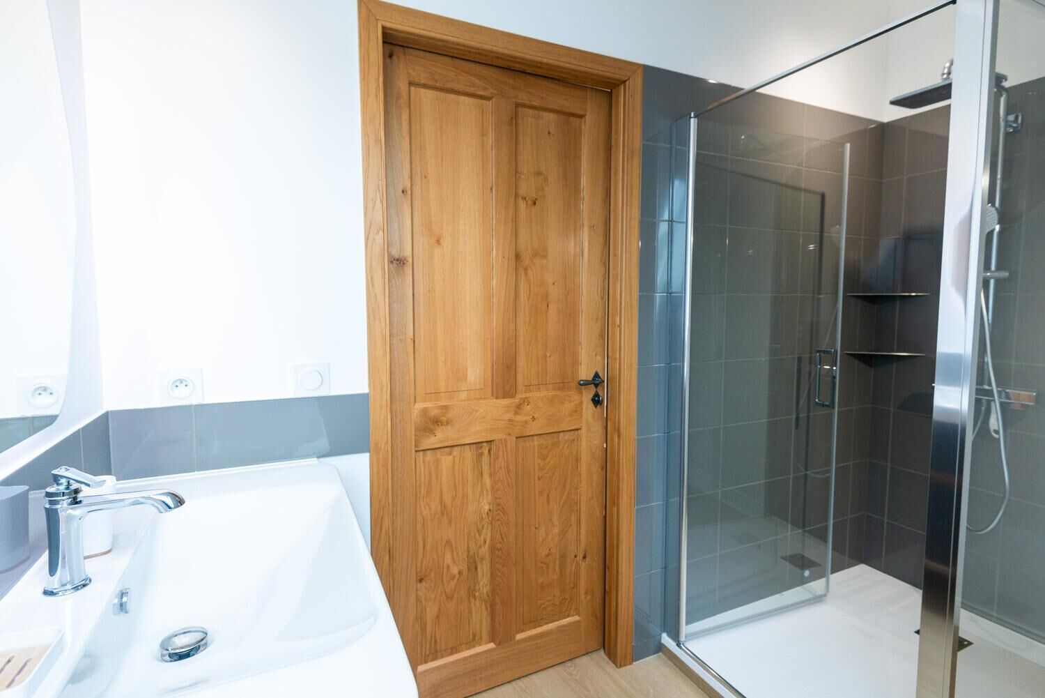 Ein Badezimmer mit Holztür, Duschkabine aus Glas und Waschbecken in Weiß.