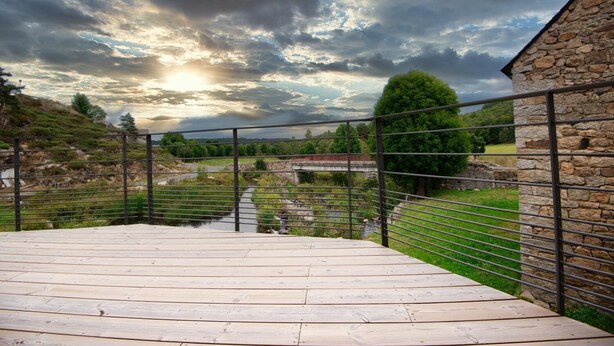A wooden deck with a metal railing overlooking a river and a stone building. - Le Moulin de Jeannou (PEYRE EN AUBRAC)