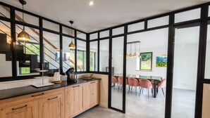 A modern kitchen with wooden cabinets, a black countertop, and a dining area with pink chairs. - Le Moulin de Jeannou (PEYRE EN AUBRAC)