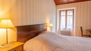 Un dormitorio con cabecera de madera, dos lámparas de noche, una colcha a cuadros, una ventana con cortinas de encaje y una mesita de noche de madera con una lámpara.