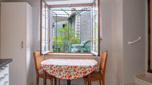 Un pequeño comedor con una mesa y dos sillas, una ventana que ofrece vistas a un jardín y un edificio, y un armario blanco a la izquierda.