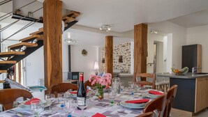Ein Essbereich mit einem Holztisch, gedeckt für vier Personen, einer Weinflasche und rosafarbenen Blumen. Im Hintergrund ist eine Küche mit einer Steinwand und einer Holztreppe zu sehen.
