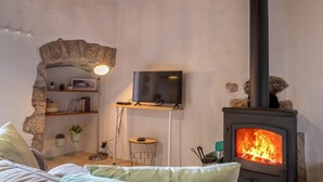 A cozy living room with a stone archway, a flat-screen TV on a wooden shelf, a round side table, and a wood-burning stove.