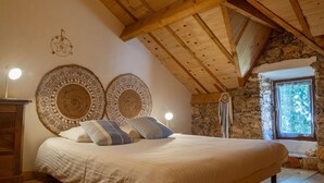 A bedroom with a large bed, wooden ceiling beams, stone walls, and two wall hangings.