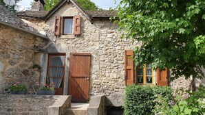 A stone house with a wooden door, a window with shutters, and a small balcony with potted plants.