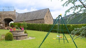Een gebouw van steen met een rieten dak, een schommel en een verzorgde tuin met potplanten en een haag.