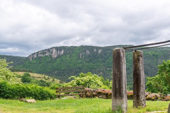 A bench in a grassy field with a stone pillar and a wooden post in the foreground, and a forested mountain in the background.