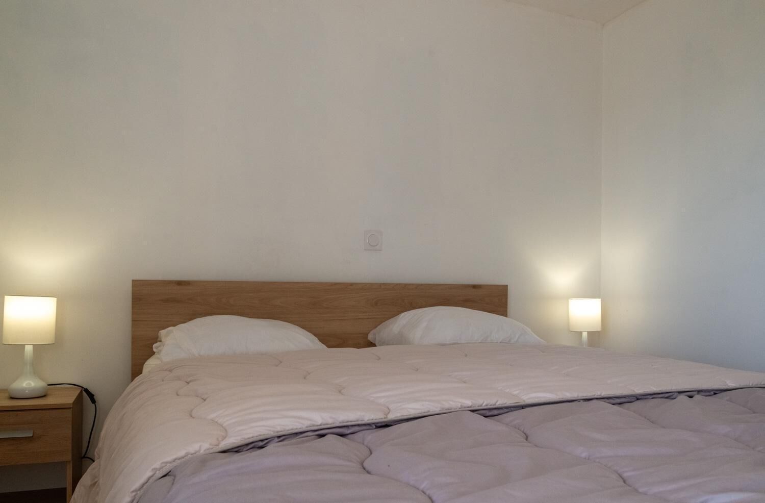 A simple, clean bedroom with a wooden headboard, two bedside lamps, and a neatly made bed.