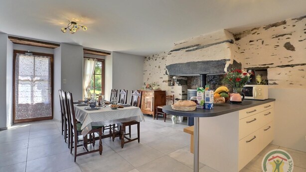A dining area with a table set for four, a kitchen with a stone wall, and a logo in the bottom right corner. - Boeuvres-corbinières (GUIPRY MESSAC)