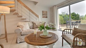 Een moderne woonkamer met een bank, een ronde houten tafel met een kaars en bloemen, een trap en een grote schuifglazen deur die uitkomt op een terras.