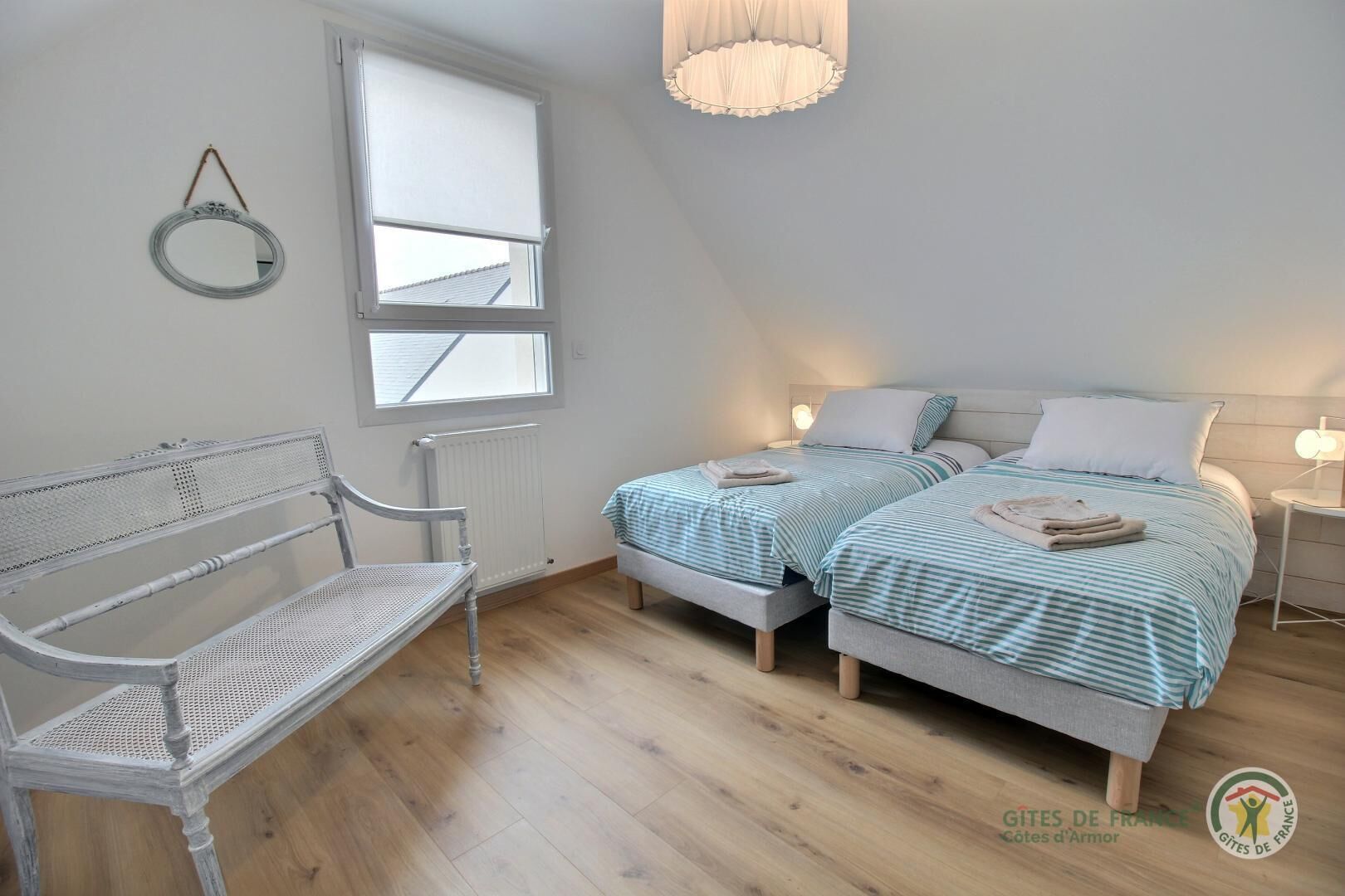 A bedroom with two beds, a bench, a window, and a hanging light fixture.