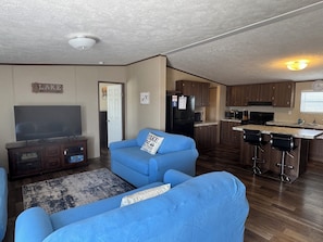 A living room with a blue sofa, a flat-screen TV, and a kitchen area with wooden cabinets and a microwave. - Spacious 4BR, 2BA, near Lake, Golf Course, and hot springs! (Elephant Butte)