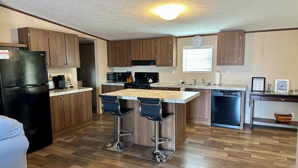 A modern kitchen with wooden cabinets, a black refrigerator, a microwave, a stove, and a sink. There are two bar stools at the island. - Spacious 4BR, 2BA, near Lake, Golf Course, and hot springs! (Elephant Butte)