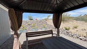 A covered outdoor seating area with a bench and a table, surrounded by pebbles and a rocky hillside with sunflowers. - Spacious 4BR, 2BA, near Lake, Golf Course, and hot springs! (Elephant Butte)