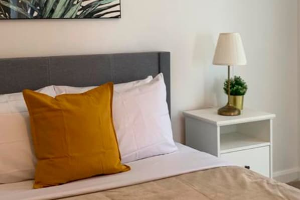 A neatly made bed with white and mustard pillows, a bedside lamp, and a small plant on a white nightstand.