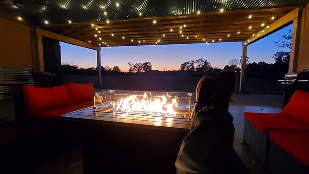 Un espacio de estar al aire libre con una mesa de vidrio y una chimenea, rodeado de cojines rojos y cubierto por una pérgola de madera con luces colgantes.