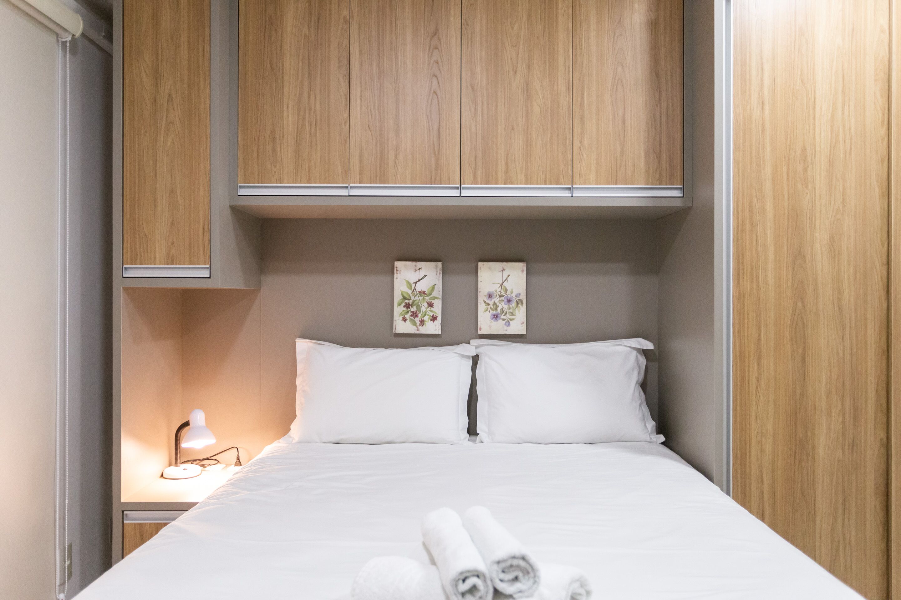 Una cama bien hecha con ropa blanca y toallas enrolladas, un armario de madera y dos cuadros de flores colgados en la pared.