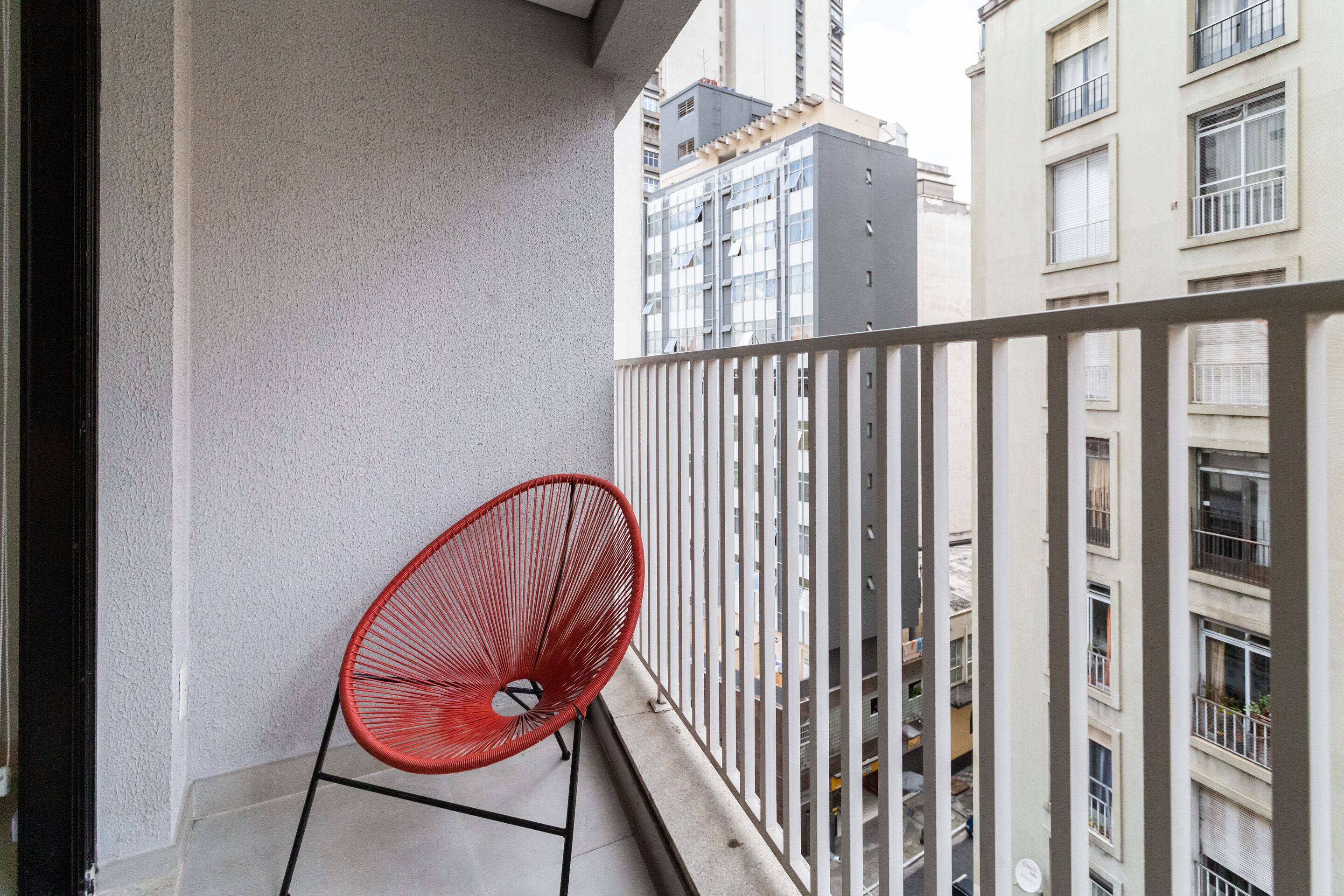 Un balcon avec une chaise rouge, une balustrade blanche et une vue sur les bâtiments voisins.