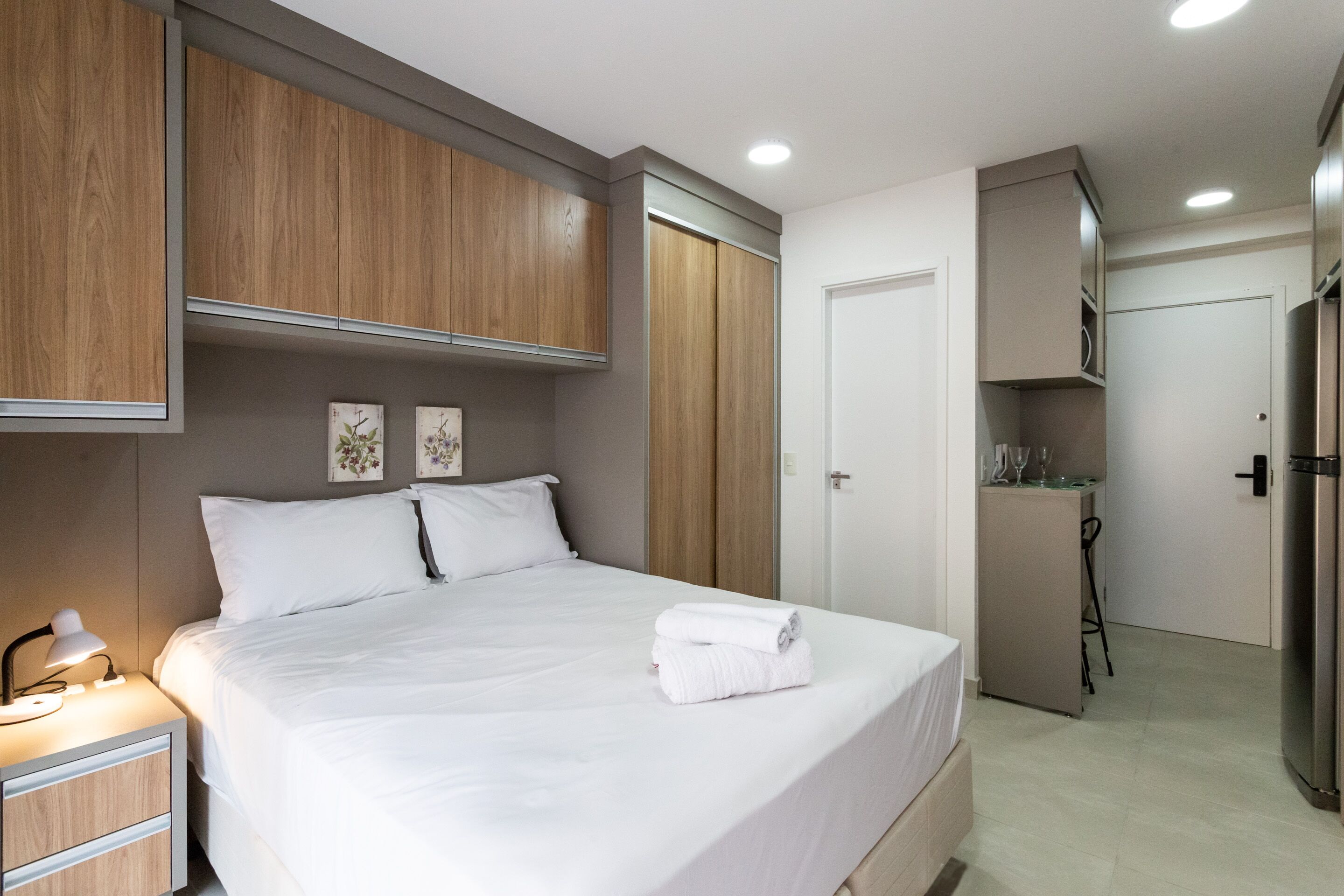 A modern hotel room with a large bed, wooden wardrobe, and a compact kitchenette.