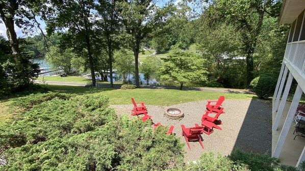 A patio with red Adirondack chairs and a fire pit surrounded by greenery, overlooking a lake.