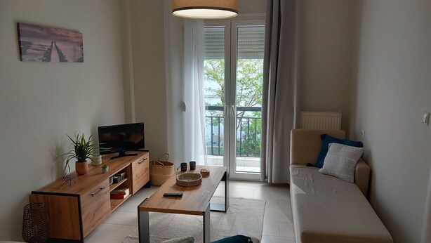 Un salon moderne avec un canapé, un téléviseur, une table d’appoint en bois et un balcon agrémenté de rideaux.