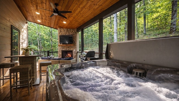 A hot tub with a stone fireplace, wicker furniture, and a view of the forest. - Hot Tub + Modern Cabin + Remote Work Setup (Broken Bow)