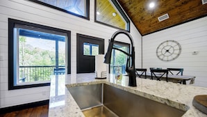 A kitchen with a stainless steel sink, wooden ceiling, and large windows offering a view of trees. - Hot Tub + Modern Cabin + Remote Work Setup (Broken Bow)