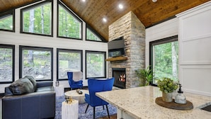 A modern interior with a stone fireplace, large windows, a seating area, and a kitchen counter. - Hot Tub + Modern Cabin + Remote Work Setup (Broken Bow)