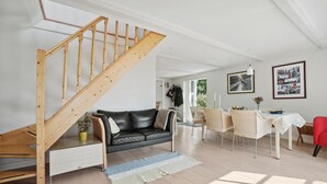 Ein modernes Wohnzimmer mit einer Holztreppe, einem schwarzen Ledersofa, einem Essbereich mit Korbstühlen und einem für zwei Personen gedecktem Tisch.