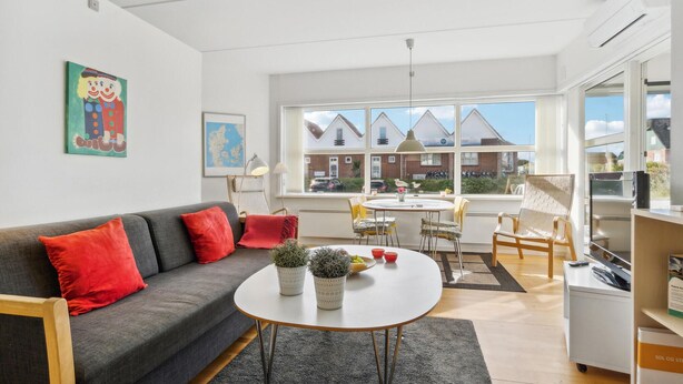Een moderne woonkamer met een grijze bank, rode kussens, een ronde salontafel met planten, een eethoek met een witte tafel en stoelen, en grote ramen met uitzicht op huizen.